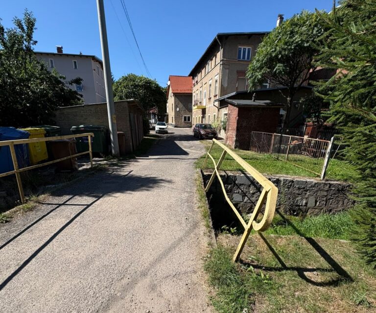 Zdjęcie przedstawia most drogowy na terenie Gminy Głuszyca przeznaczony do odbudowy. Widoczna jest wąska asfaltowa jezdnia przebiegająca przez most, po obu stronach zabezpieczona żółtymi metalowymi barierkami. W otoczeniu znajdują się drzewa, krzewy oraz zabudowania.