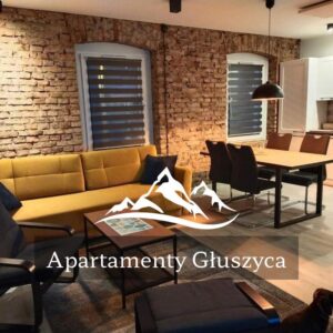 Przestronne, nowoczesne wnętrze Apartamentów Głuszyca z częścią wypoczynkową, jadalnią i aneksem kuchennym. Charakteru dodaje mu oryginalna ceglana ściana oraz ciepłe oświetlenie. Na pierwszym planie widoczna jest wygodna sofa i fotele, a w głębi stół z krzesłami. Na zdjęciu znajduje się logo i napis „Apartamenty Głuszyca”.