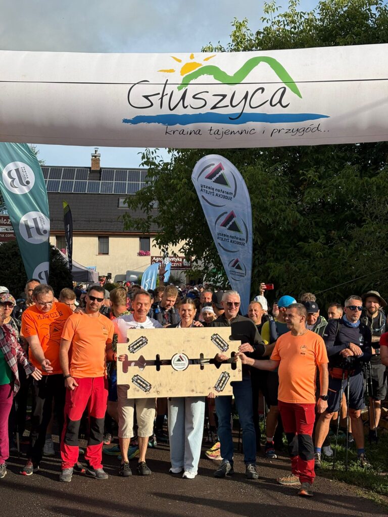 Uczestnicy i organizatorzy 19. Maratonu Pieszego „Sudecka Żyleta” zgromadzeni na starcie w Głuszycy Górnej. Na pierwszym planie grupa osób trzyma dużą drewnianą tablicę w kształcie żyletki, w tle zawodnicy przygotowani do wyruszenia na trasę.