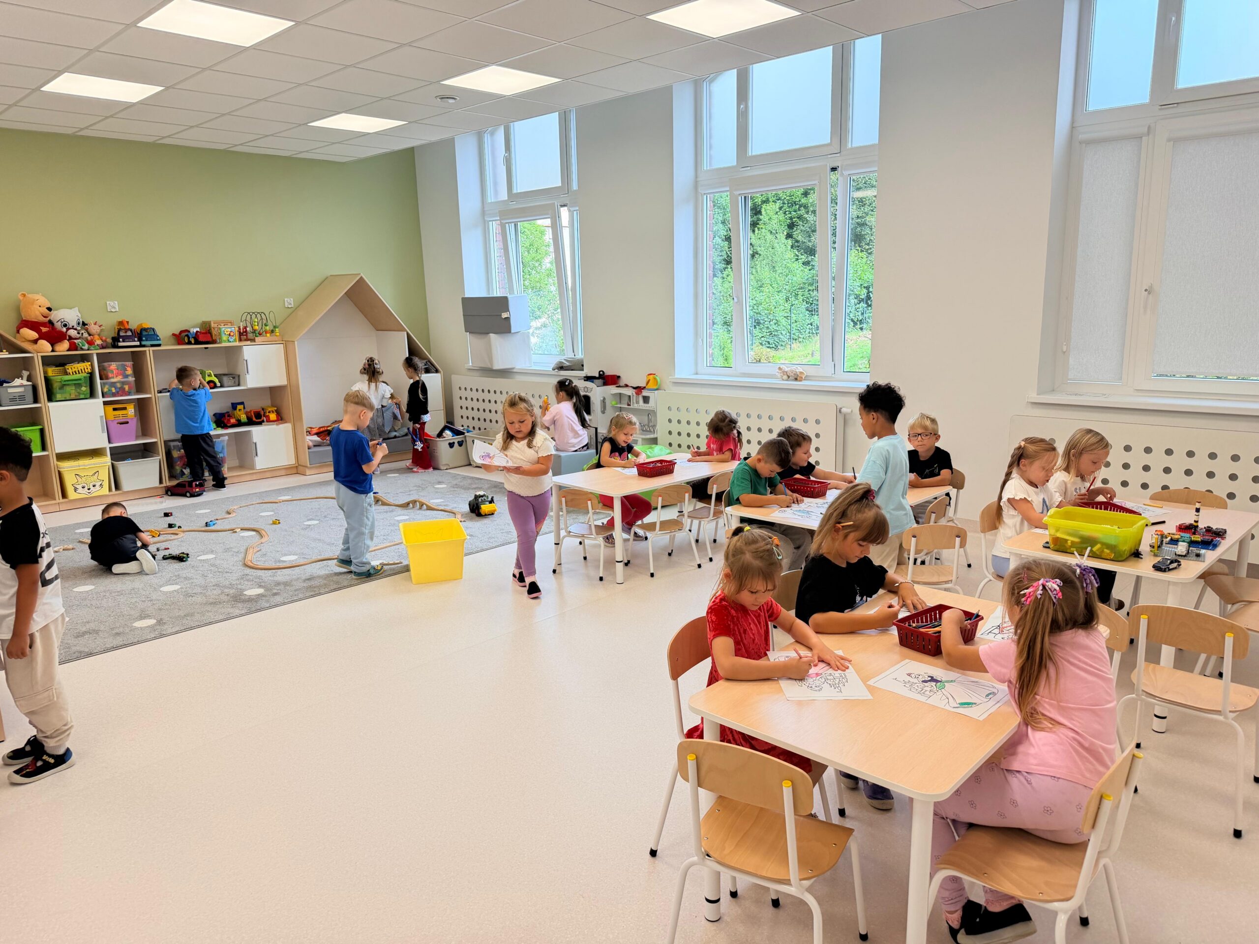 Jasna, przestronna sala przedszkolna. Kilkanaścioro dzieci uczestniczy w zajęciach – część siedzi przy stolikach i koloruje obrazki, a pozostałe bawią się zabawkami oraz drewnianą kolejką na dywanie. W sali znajdują się niskie regały z zabawkami, małe stoliki i krzesła oraz duże okna.